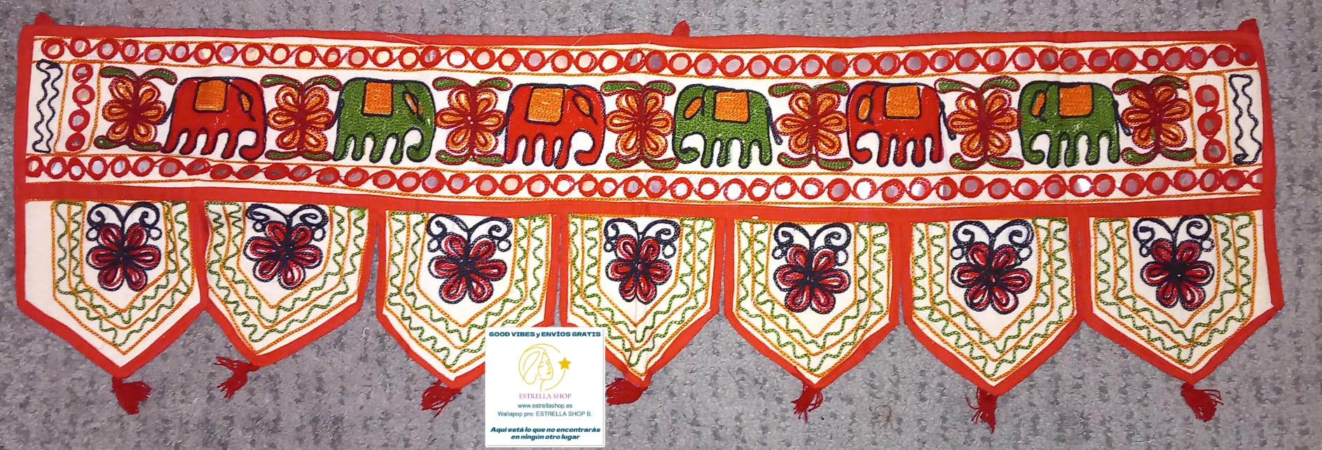 Un detalle que transforma cualquier espacio con esencia étnica y espiritual. El Torán Gora de 30x100 cm es una pieza elaborada artesanalmente en algodón con fondo color hueso, decorada con bordados coloridos y pequeños espejos que capturan la luz. Tradicionalmente usado en India para decorar puertas como símbolo de bienvenida y protección, hoy es ideal para paredes, escaparates, mostradores o como toque original en stands de mercado. Cada unidad es única, con composiciones distintas que harán que tu ambient