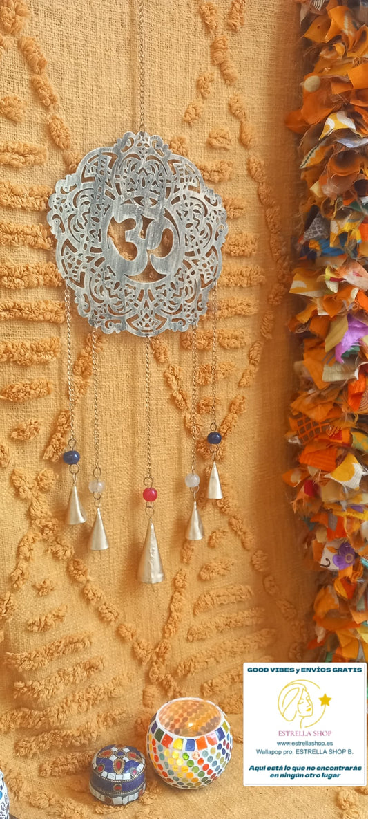 Un objeto decorativo con simbolismo profundo y belleza artesanal. El colgador metálico Atrapasueños con Om, elaborado en tono dorado apagado, une dos poderosos símbolos de la espiritualidad india: el mantra Om. Forma redondeada desde donde cuelgan cinco cadenas de diferentes longitudes decoradas con abalorios de cristal y terminaciones con campanas.Con una longitud aproximada de 67 cm y 22 cm de diámetro, este colgador no solo embellece cualquier espacio, sino que también genera un suave y relajante tintine