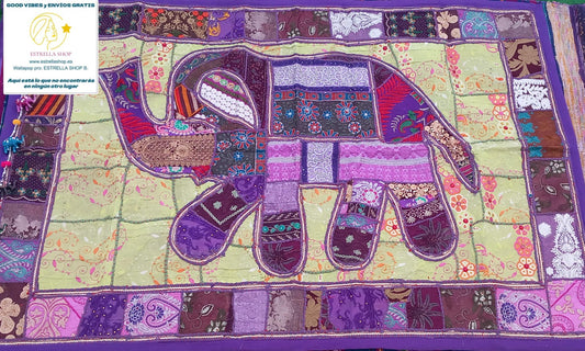 Un clásico del arte textil reinventado con un toque exótico y exclusivo. El tapiz patchwork cuadrado de 150x100 cm está confeccionado artesanalmente con retales bordados en algodón, creando composiciones únicas con flores, formas geométricas y pequeños abalorios brillantes. Cada pieza tiene su propio carácter, con un ribete en tono coordinado que enmarca el diseño y lo convierte en una joya decorativa. Ideal para colgar, usar como camino de mesa o sobre un mueble que quieras destacar.